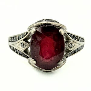 Puede incluir: Un anillo de plata con una gran piedra preciosa roja oscura de corte ovalado como pieza central. La banda está adornada con piedras preciosas negras y detalles plateados, creando un contraste llamativo. El diseño del anillo es intrincado y elegante.