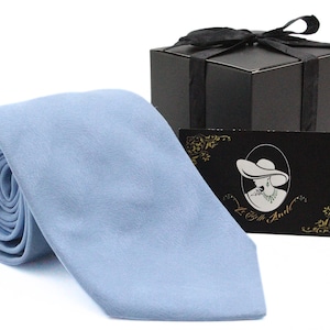 May include: A rolled-up, light blue tie with a smooth texture. A black gift box with a black ribbon and a business card with a lady illustration are behind the tie. The business card has gold accents.