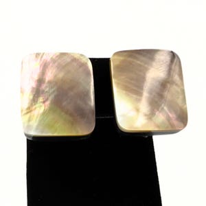 May include: Pair of iridescent square earrings. The earrings have a mother-of-pearl appearance with a mix of colors, including pink, green, and gold. The earrings are displayed on a black surface against a white background.