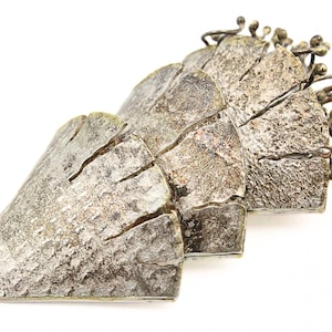 Könnte beinhalten: Eine silberfarbene Metallskulptur mit strukturierter Oberfläche und geschichtetem Design. Die Skulptur hat eine Reihe von sich überlappenden, eckigen Formen, mit einer Ansammlung kleiner, astähnlicher Elemente, die sich von oben erstrecken. Auf weißem Hintergrund.