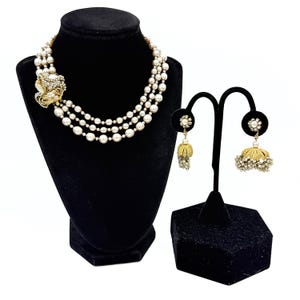 Pode incluir: Um colar de pérolas de três fios com um detalhe lateral dourado com cristais transparentes e um par de brincos jhumka dourados a condizer com detalhes de pérolas e tachas de cristal, expostos em suportes de joalharia de veludo preto.