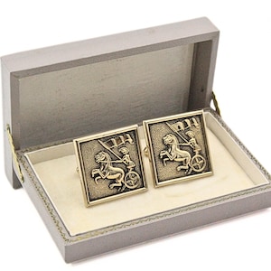 May include: A pair of gold-toned cufflinks featuring a chariot scene with a horse and figure, presented in a gray box with a cream-colored interior. The cufflinks are square-shaped and have a detailed design.