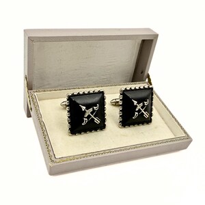 May include: A pair of square cufflinks with a black face and silver trim, presented in a gray box. Each cufflink features a silver design of crossed arrows. The box is open, revealing a cream-colored interior.