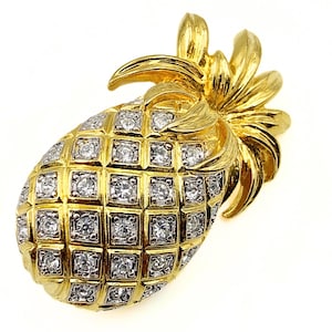 Op de afbeelding: Een goudkleurige ananasbroche met een gestructureerde metalen afwerking en een ruitpatroon. Elk vierkant gedeelte van het ananaslichaam is ingelegd met een kleine, heldere, ronde strass-steen, terwijl de bovenste bladeren van glad goudmetaal zijn.