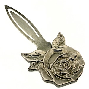 Peut inclure: Un marque-page en métal argenté orné d'une rose en relief détaillée avec des feuilles à la base. La partie supérieure du marque-page présente une forme ovale allongée et fine avec une découpe centrale, s'étendant depuis l'ornement floral.