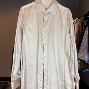 May include: A long-sleeved, button-down shirt in a light beige color. The shirt has a classic collar and cuffs, and is hanging on a wooden rack. The fabric appears to have a slight sheen.
