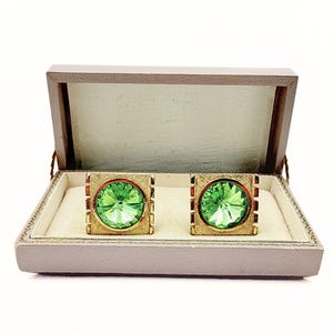 May include: A pair of square gold-tone cufflinks featuring a central round, faceted light green stone. The metal surface has a textured, brushed finish with vertical grooved detailing on the sides, presented in a rectangular hinged box.