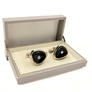 May include: A pair of gold-toned cufflinks featuring a triangular shape with a smooth black inlay and a single clear stone set in the center. The cufflinks are presented inside a rectangular, light gray hinged box with a cream-colored interior.