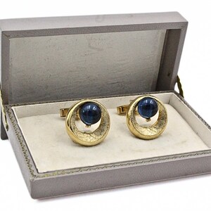 May include: A pair of gold-toned cufflinks with a blue orb center, presented in a gray box with a cream-colored interior. The cufflinks have a circular design with a textured pattern.