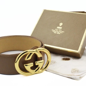 May include: A brown leather belt with a gold-colored buckle featuring the interlocking "G" logo. The belt is displayed with its original box and a protective cloth. The box has gold trim and the brand name.