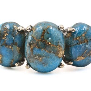 Könnte beinhalten: Ein silbernes Armband mit drei ovalen, blau-grünen Steinen mit goldfarbenen Adern. Die Steine sind in silbernen Zinken gefasst, vor weißem Hintergrund.