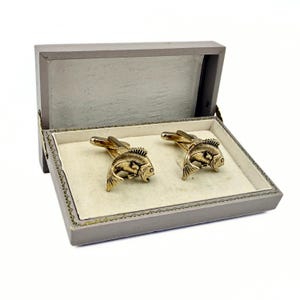 May include: A pair of gold-toned fish cufflinks displayed in a gray box with a cream-colored interior. The cufflinks are detailed with scales and fins. The box is open, revealing the cufflinks.