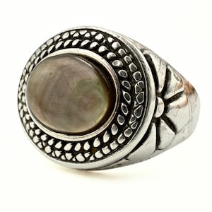 May include: A silver ring featuring a large, oval, iridescent grey stone. The ring has a detailed, textured design around the stone and on the band, with a pattern of small beads and floral elements. A statement ring.