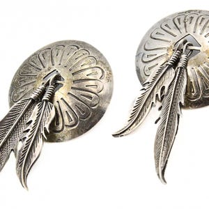 Könnte beinhalten: Ein Paar silberne Ohrringe mit einem runden Design und einem Federnanhänger. Die Ohrringe sind aus Sterlingsilber gefertigt und haben ein indianisch inspiriertes Design.