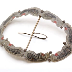 May include: An oval-shaped metal belt buckle with a paisley design. The buckle is decorated with small rhinestones and has a red and black color scheme.