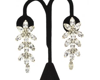 May include: A pair of sparkling, clear crystal earrings on a black display stand. Each earring features a floral cluster at the top, with cascading crystal leaves and a single teardrop crystal at the bottom. The earrings are designed to dangle.