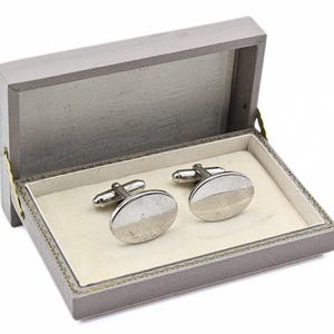 May include: Silver oval cufflinks in a grey presentation box. The cufflinks have a two-tone design. The box has a cream-coloured interior. The cufflinks are a classic accessory.