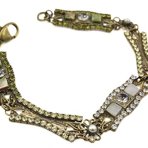 May include: A gold-tone bracelet with a geometric design featuring green and white stones. The bracelet has a clasp closure.