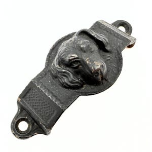 Könnte beinhalten: Ein dekoratives Stück aus schwarzem Metall in Form eines Hundekopfes. Der Hundekopf befindet sich in der Mitte des Stücks, mit einem rechteckigen Abschnitt auf beiden Seiten. Das Stück hat an einem Ende eine Schlaufe und am anderen ein strukturiertes Muster.