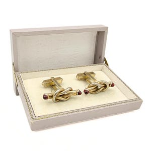 May include: A pair of gold-toned metal cufflinks featuring a knotted design with small, dark red cabochon accents at the ends. The cufflinks are presented inside a rectangular, light gray hinged storage box with a cream-colored interior lining.