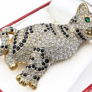 May include: A bejeweled cat brooch with clear and black crystals, gold accents, and green eyes. The cat is posed lying down, with its tail curled. The brooch is presented in a red and white box.