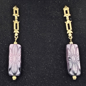 May include: A pair of gold-toned earrings with a rectangular, lavender-colored pendant. The pendant features a carved floral design. The earrings have a decorative top and a small gold bead at the bottom of the pendant.