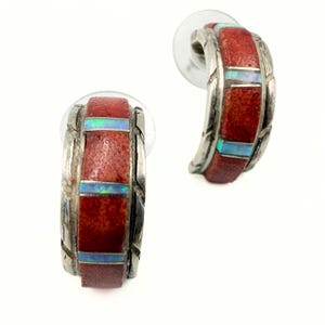 May include: A pair of silver hoop earrings with a red coral center and inlaid opal accents. The earrings have a textured design on the silver and are shown against a white background. The earrings are a unique and stylish accessory.