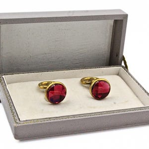 May include: A pair of round cufflinks featuring faceted deep red stones set in gold-toned metal, presented inside a rectangular gray hinged storage box with a white interior lining. The cufflinks have a circular face and a gold-toned loop backing.
