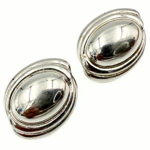 May include: A pair of silver-toned oval clip-on earrings. Each earring has a smooth, reflective centre surrounded by raised, ridged bands. The earrings are set against a bright white background, highlighting their polished surface.