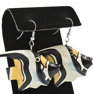 Puede incluir: Un par de pendientes hechos a mano con un diseño de pez. Los pendientes están pintados con detalles en negro, blanco y amarillo. Los pendientes con forma de pez cuelgan de ganchos plateados, expuestos en un expositor de joyas negro.