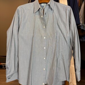 Peut inclure: Une chemise boutonnée à carreaux gris et blancs avec des manches longues. La chemise est suspendue à un cintre en bois. La chemise a un col classique et des poignets boutonnés. La chemise est en tissu léger.