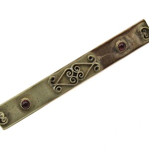 May include: A silver-toned barrette with ornate filigree and three small, round, red gemstones. The barrette features decorative scrollwork and is set against a white background. The barrette is approximately 4 inches long.