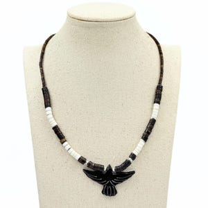 May include: A necklace with a black bird pendant. The necklace has a brown beaded strand with white and dark brown accents. The pendant is carved and has a glossy finish. The necklace is displayed on a mannequin. The necklace is approximately 45 cm long.