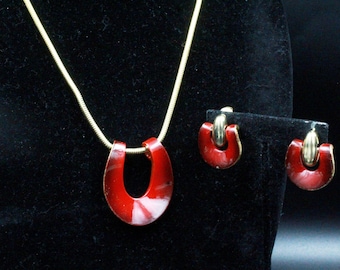 Puede incluir: Un conjunto de collar con colgante rojo y pendientes a juego. El colgante y los pendientes tienen un acento de metal dorado. La cadena del collar es una fina cadena de serpiente dorada. Las joyas se exhiben sobre una superficie negra.