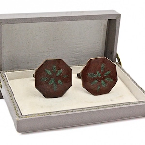 May include: A pair of octagonal cufflinks featuring a deep maroon enamel finish with a central green floral pattern, presented inside a rectangular grey storage box with a white interior lining and a hinged lid that opens to display the items.