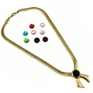 Puede incluir: Un collar dorado con una cadena de espiga y un colgante en forma de cruz con una piedra negra. La imagen también muestra una colección de cuentas redondas de colores en rojo, blanco, azul, rosa, plateado, negro y marrón.