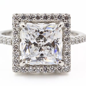 Op de afbeelding: Een zilveren ring met een grote vierkante diamant omgeven door een halo van kleinere diamanten.