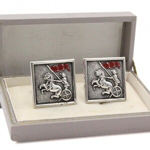 May include: Silver-toned cufflinks with a detailed design of a person in a chariot pulled by a horse, holding a red flag. The cufflinks are square and presented in a gray box with a cream-colored interior.