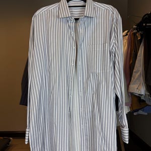 May include: A grey and white striped long-sleeved shirt hanging on a black hanger. The shirt features a button-down front, a chest pocket, and buttoned cuffs. The shirt is made of a lightweight fabric.
