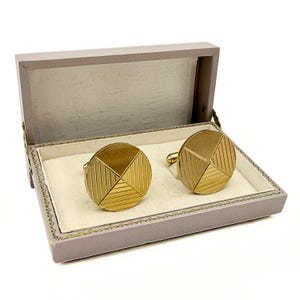 May include: A pair of gold-toned cufflinks with a geometric design, presented in a gray box with a light-colored interior. The cufflinks have a circular shape, divided into sections with textured lines.