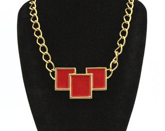 Puede incluir: Collar de cadena dorada con tres colgantes cuadrados rojos. El collar se exhibe en un maniquí negro, resaltando el contraste de color. La cadena tiene un diseño de eslabones gruesos.