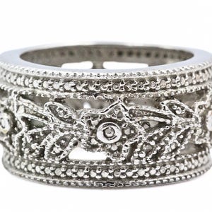 Könnte beinhalten: Ein silberfarbener Ring mit einem aufwendigen Blumendesign. Der Ring weist ein detailliertes Muster aus Blumen und Blättern mit einer strukturierten Oberfläche auf. Der Ring ist im Bandstil gehalten, mit einem durchgehenden Design.
