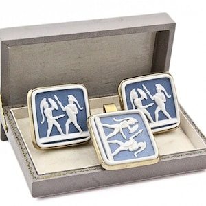 May include: A set of cufflinks and a tie clip in a gray box. The square cufflinks and tie clip feature a white relief design on a blue background, framed in gold. The relief depicts two figures.