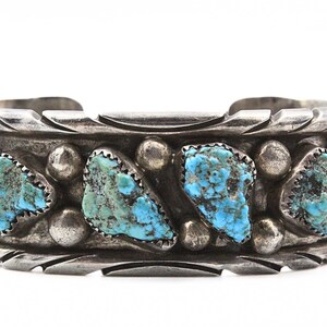 May include: A silver cuff bracelet with turquoise stones set in a geometric pattern. The bracelet has a textured design and is made of sterling silver.