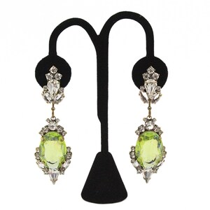 Pode incluir: Um par de brincos pendurados ornamentados. Cada brinco apresenta uma grande pedra oval verde claro, rodeada por cristais transparentes e brilhantes. Os brincos são exibidos em um suporte de veludo preto, mostrando seu design intrincado e estilo elegante.
