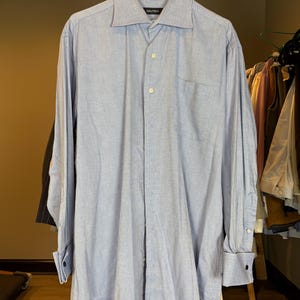 May include: Light blue long-sleeve shirt with a button-down front, a chest pocket, and a classic collar. The shirt is branded with the "NAUTICA" logo on the collar. The shirt is hanging on a wooden rack.