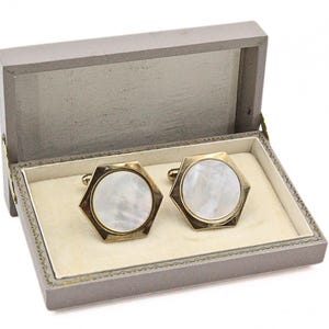 May include: A pair of gold-toned, hexagonal cufflinks with mother-of-pearl centers, presented in a gray, hinged box with a cream-colored interior. The box is open, displaying the cufflinks.