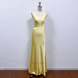 Puede incluir: Un vestido largo de color amarillo pálido con escote en V y tirantes finos. El vestido está hecho de un material sedoso y tiene un corpiño ajustado y una falda fluida. El vestido se exhibe en un maniquí. El texto "LAUSEN CRAFTS" es visible.