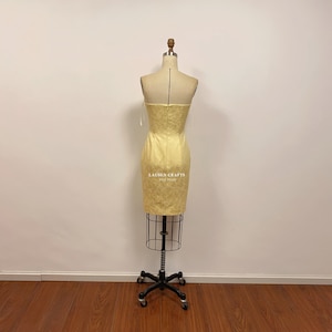 May include: A yellow strapless dress with a fitted silhouette and a textured fabric. The dress has a small bow detail on each shoulder strap. The dress is on a mannequin and the background is a white wall.