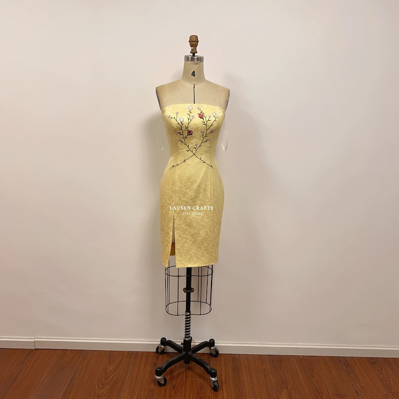 May include: A yellow strapless dress with a floral embroidery design. The dress has a fitted silhouette and a slit on the side. The dress is displayed on a mannequin.
